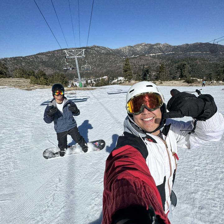 Matt taking a selfie on a snowy slope with a friend on a snowboard behind him